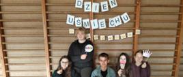 Pięcioro uczniów pozuje do zdjęcia z papierowymi gadżetami. Za nimi na drabinach widnieje napis DZIEŃ UŚMIECHU oraz obrazki przedstawiające uśmiechnięte buźki.
