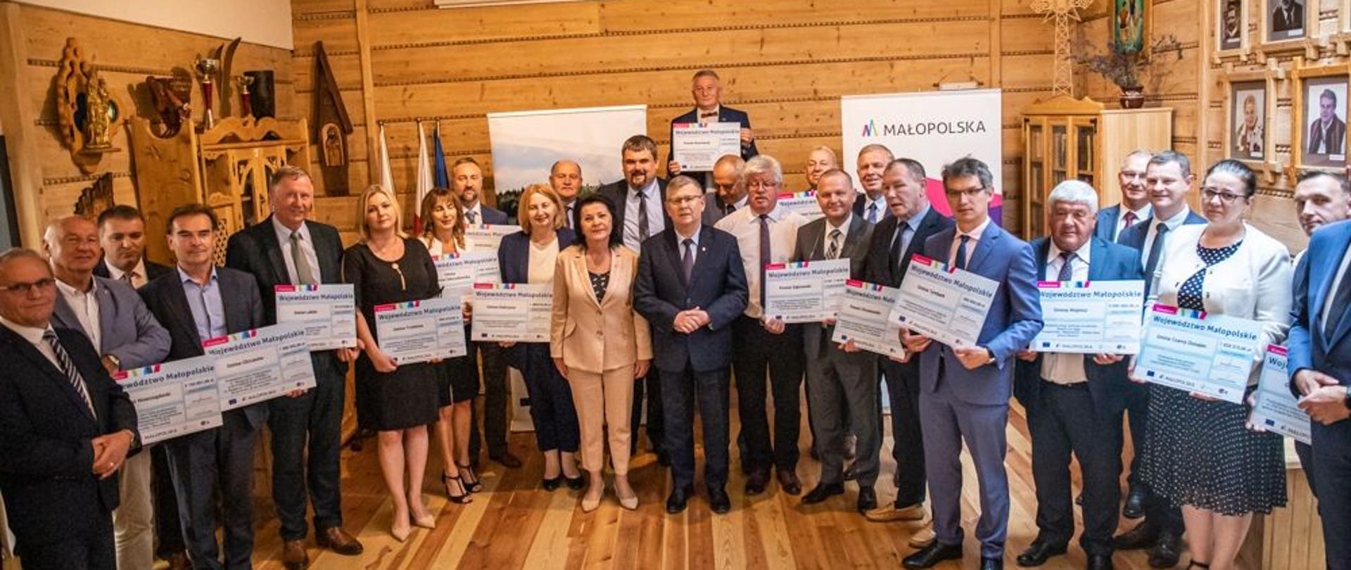 Zdjęcie grupowe: Wójtowie i burmistrzowe trzymający symboliczne promesy. w centralnej części zdjęcia marszałek Małopolski Witold Kozłowski, senator Jan Hamerski, poseł Anna Paluch, poseł Patryk Wicher oraz radny województwa Stanisław Barnaś