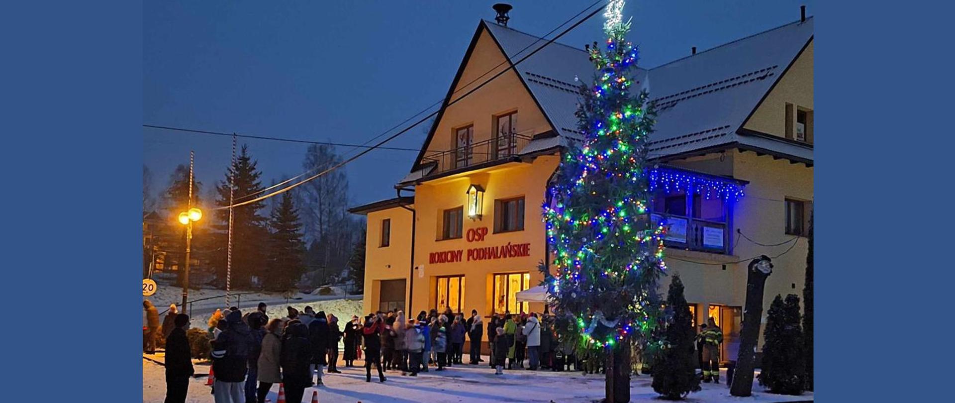 Świąteczna choinka stojąca na placu przy remizie OSP wystrojona w setki światełek wraz przybyłymi mieszkańcami .