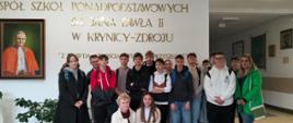 Cała klasa ósma stoi w grupie w holu Zespołu Szkół Ponadpodstawowych w krynicy-Zdroju, na ścianie obraz przedstawiający papieża Jan Pawła II