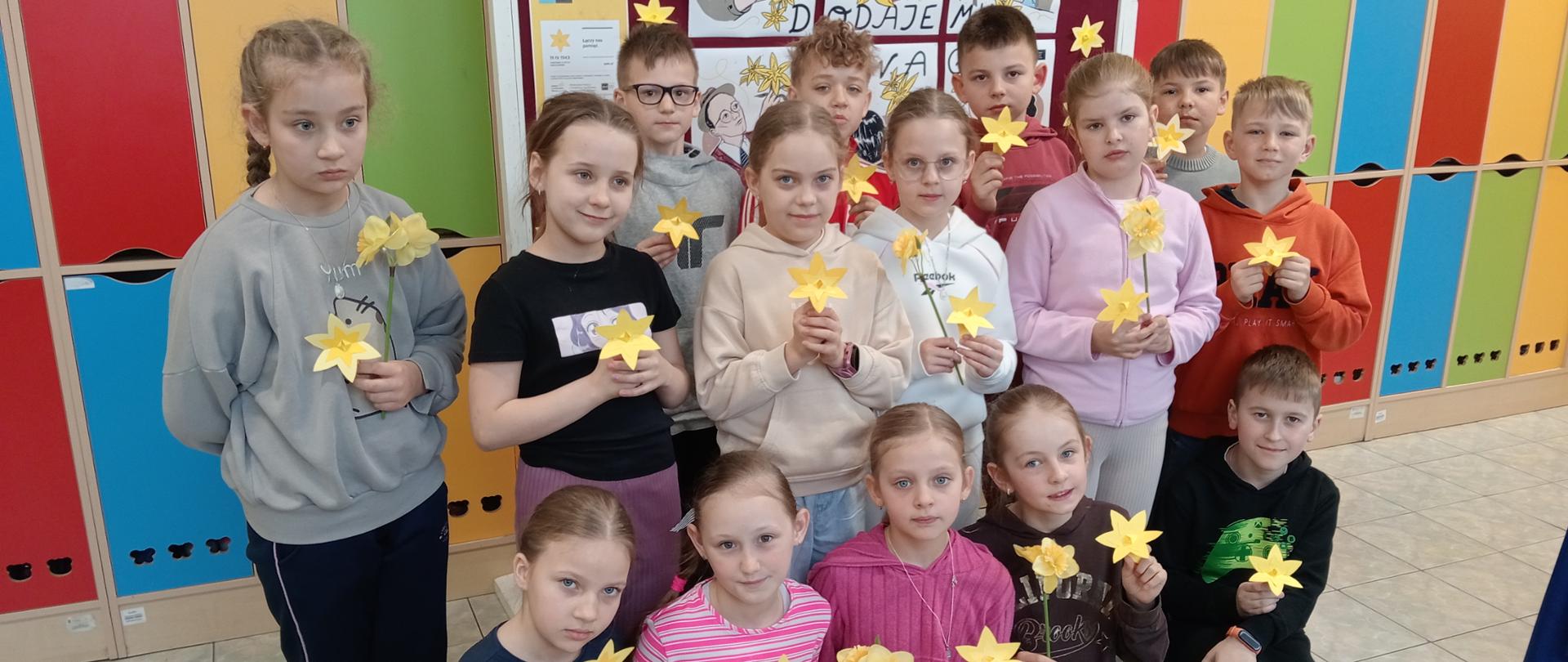 Przed tablicą, na której znajdują się plakaty związane z akcją, stoją dzieci trzymające w rękach papierowe żonkile