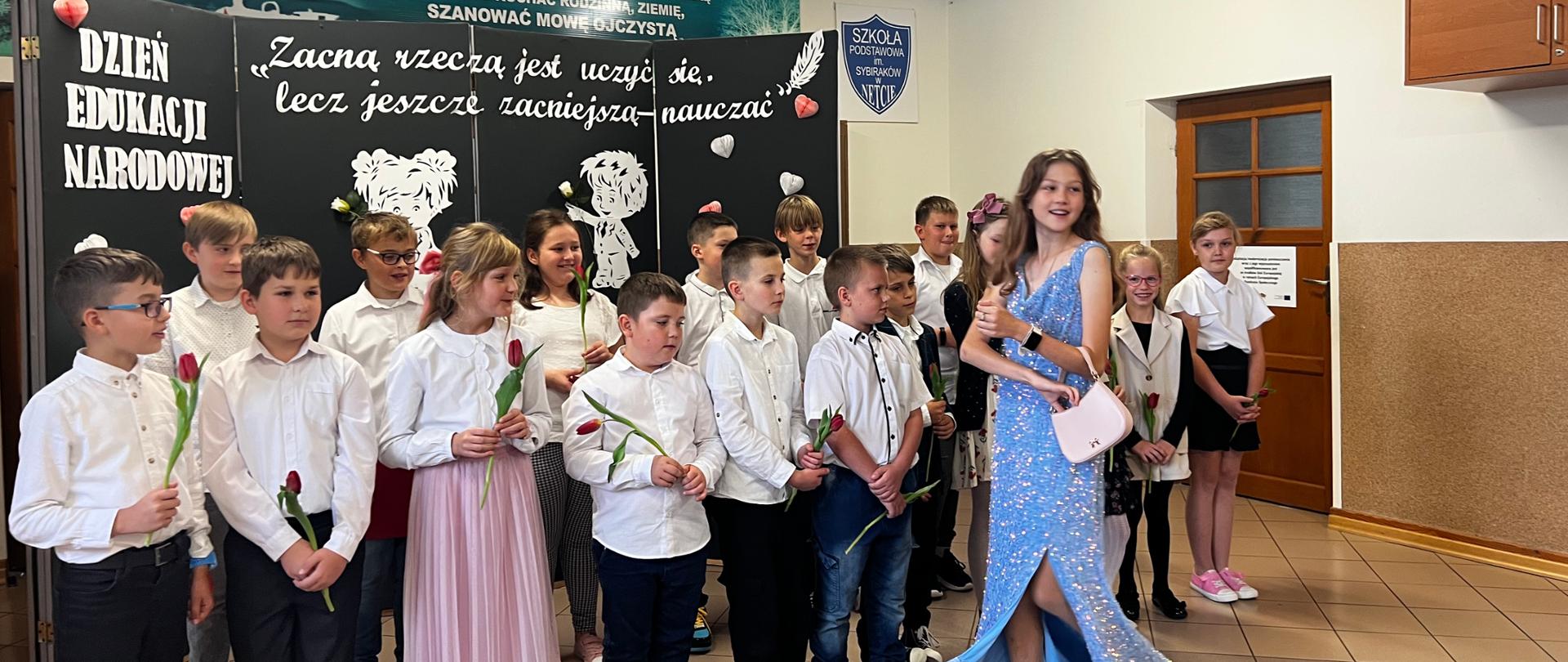 Na środku znajduje się grupa uczniów ubranych w stroje galowe. Przed nimi przechadza się dziewczyna ubrana w długą, niebieską suknię balową. Uczennica uśmiecha się w stronę widowni. Za nimi, na ściance widnieje napis: DZIEŃ EDUKACJI NARODOWEJ. Zacną rzeczą jest uczyć się, lecz jeszcze zacniejszą - nauczać.