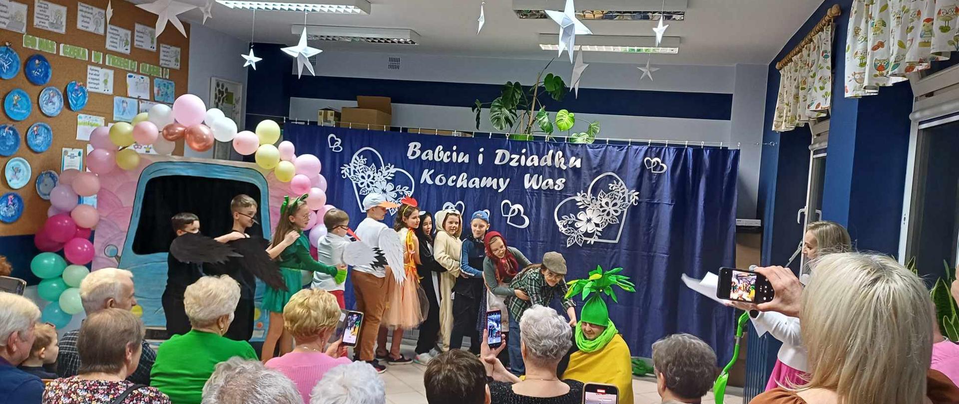 Grupa dzieci w przebraniach trzymająca się za ramiona i biodra. Pierwsza osoba ciągnie chłopca w zielonej czapce z liściami. Przed nimi siedzą starsi ludzie.