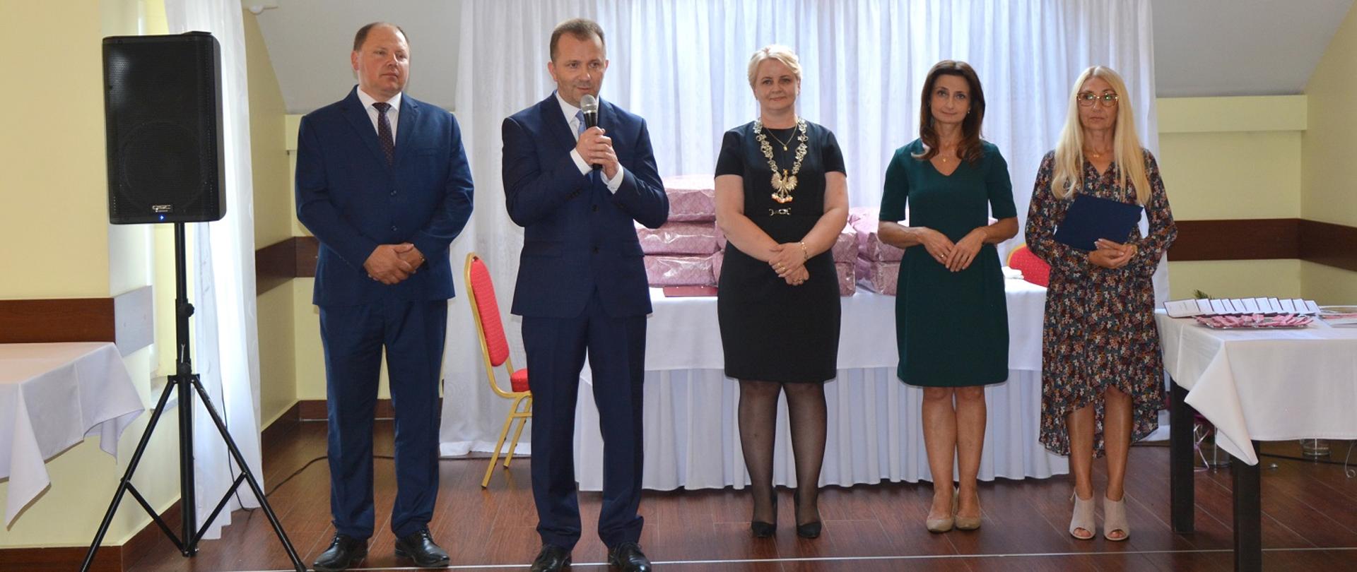 dwóch mężczyzn z ciemnych garniturach i trzy kobiety w eleganckich sukienkach stoją w rzędzie, jeden z mężczyzna przemawia do mikrofonu 