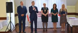 dwóch mężczyzn z ciemnych garniturach i trzy kobiety w eleganckich sukienkach stoją w rzędzie, jeden z mężczyzna przemawia do mikrofonu 