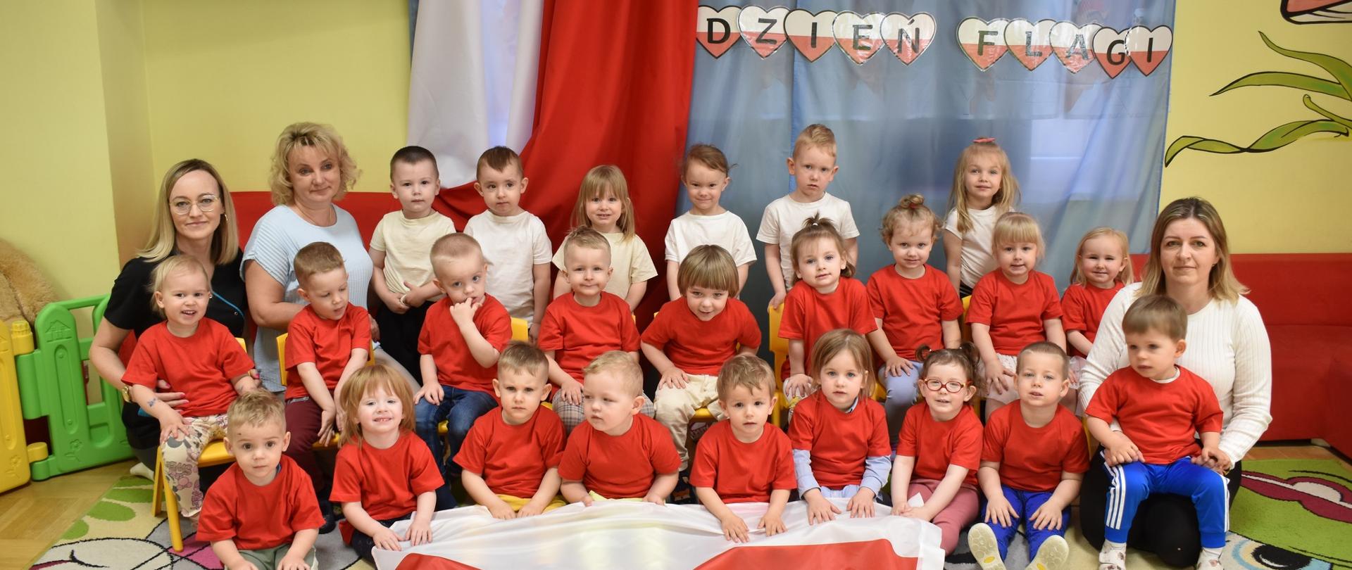 Na zdjęciu widać grupę dzieci, ustawione w trzech rzędachi ubrane w odświętne biało-granatowe stroje oraz panie opiekunki z Panią Dyrektor. Na tle widnieje napis dzień Flagi