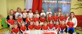 Na zdjęciu widać grupę dzieci, ustawione w trzech rzędachi ubrane w odświętne biało-granatowe stroje oraz panie opiekunki z Panią Dyrektor. Na tle widnieje napis dzień Flagi