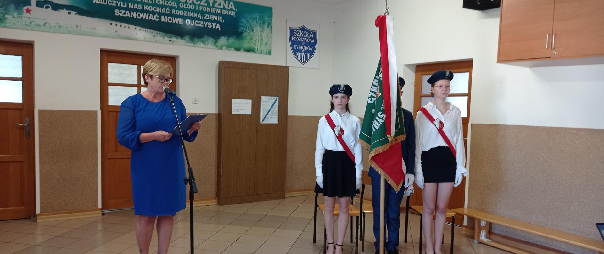 Pani Dyrektor odczytuje przez mikrofon treść kartki, którą trzyma w ręce. Ubrana jest w niebieską sukienkę. Obok stoją ubrani na galowo uczniowie z pocztem sztandarowym.