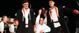 Dwóch mężczyzn w ciemnych spodniach, białych koszulach i czarnych marynarkach niosą wiklinowy kosz w którym siedzi mężczyzna w ciemnych spodniach i białej koszuli 