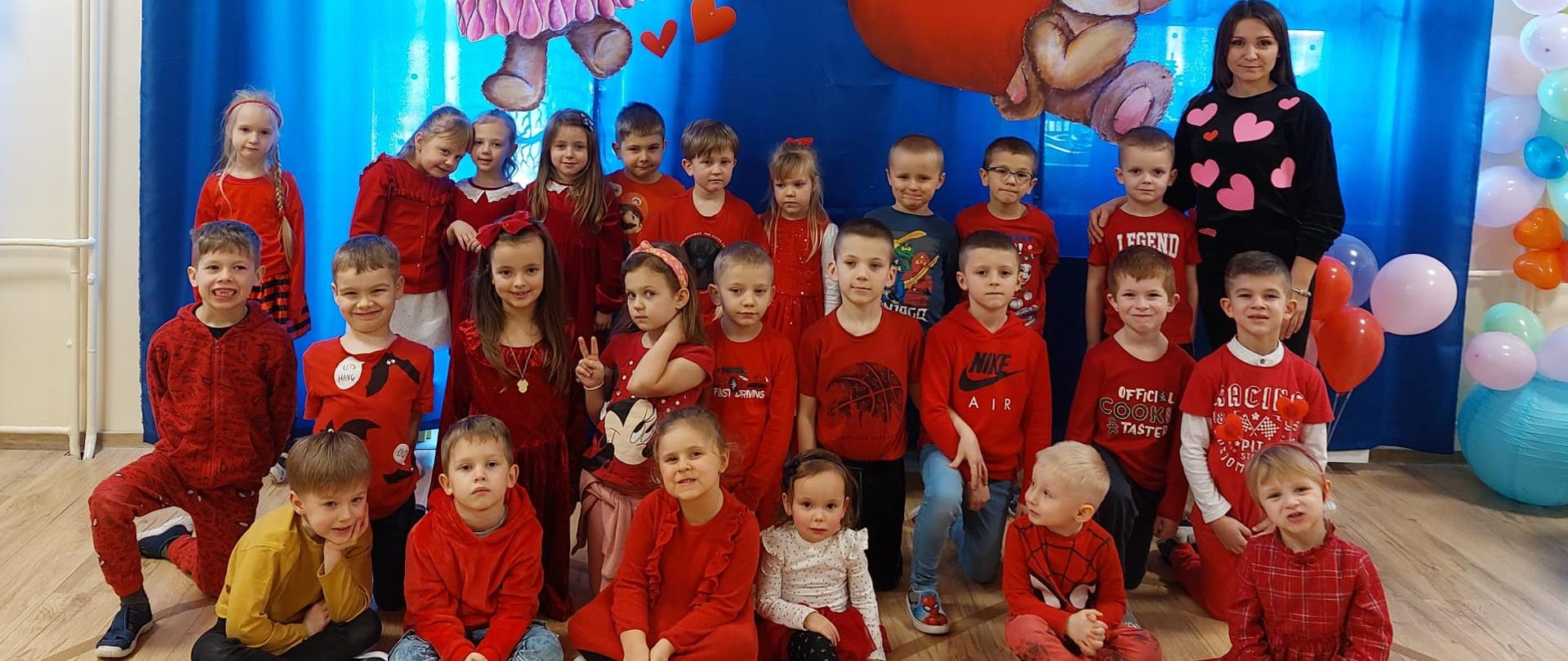 Zdjęcie grupowe przedszkolaków ubranych na czerwono, wraz z wychowawczynią, na tle walentynkowej dekoracji. 