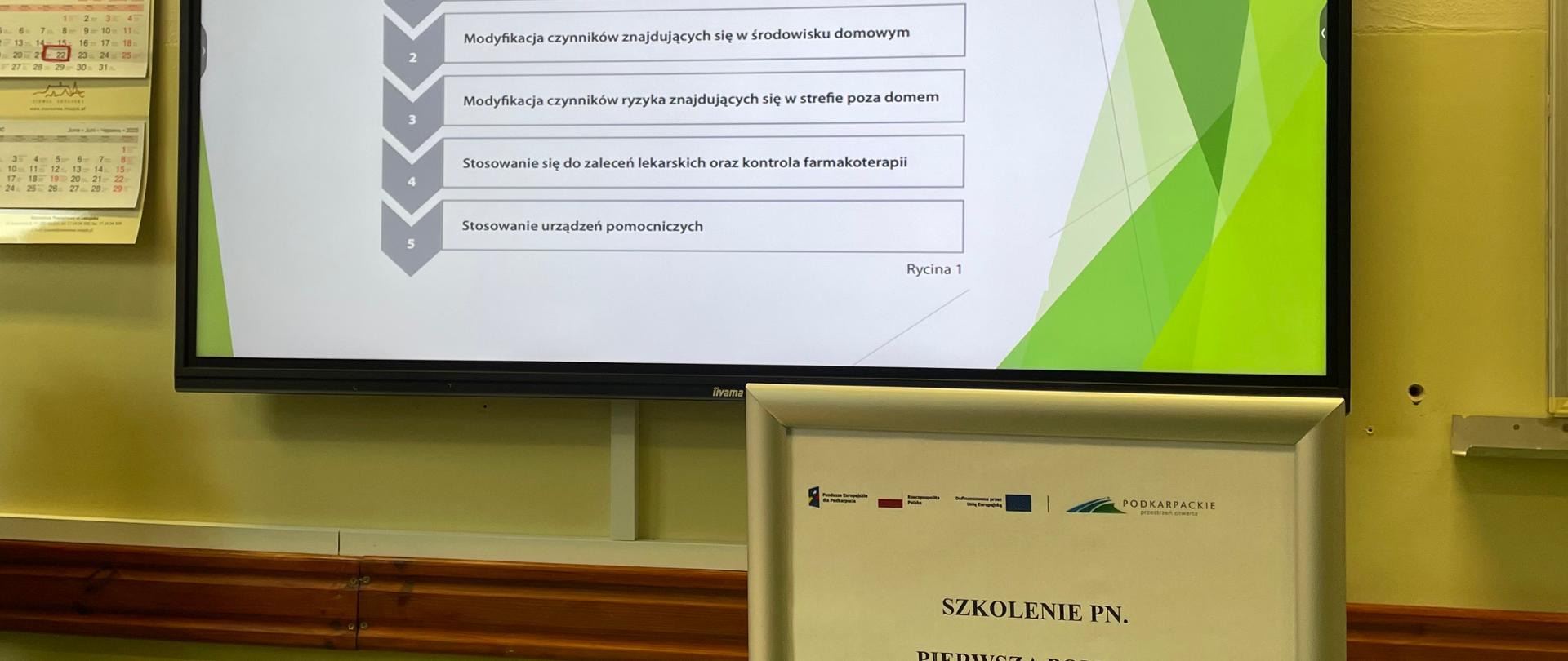 Rysunek przedstawia tablicę na której wyświetlona jest treść szkolenia, przed którą znaduje się statyw z napisem szkolenie i innymi treściami. 