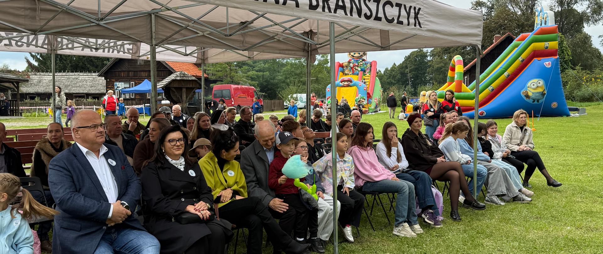 Pierwsze rzędy widowni siedzące w cieniu giętego namiotu gminy Brańszczyk. Siedzący ludzie, w tym dorosły mężczyzna w garniturze oraz kobiety i dzieci, obserwują wydarzenia na scenie.