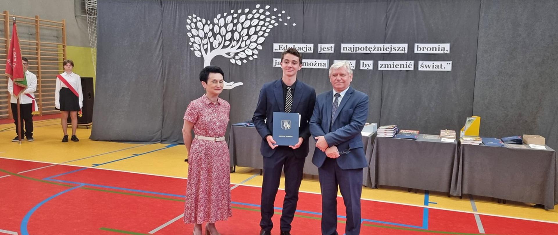 Wójt, Dyrektor Szkoły oraz nagrodzony uczeń stoją tle szarej dekoracji. Z tyłu dwoje uczniów ze sztandarem szkoły.