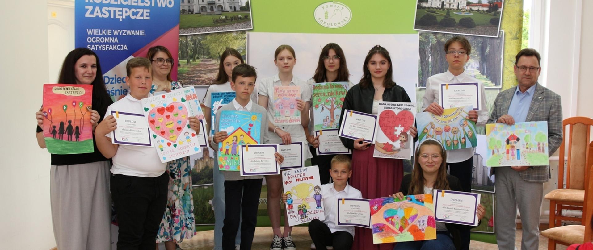 Fotografia grupowa laureatów w konkursie plastycznym. Rozstrzygnięcie odbyło się w sali konferencyjnej Starostwa Powiatowego w Sokołowie Podlaskim. Od lewej dyrektor PCPR oraz uczestnicy i opiekunowie, po prawej stronie starosta sokołowski. Wszyscy prezentują w dłoniach prace plastyczne. Za nimi ścianka powiatu sokołowskiego z logo, napisem powiat sokołowski i fotografiami z terenu powiatu oraz roll - up z napisem Rodzicielstwo zastępcze.