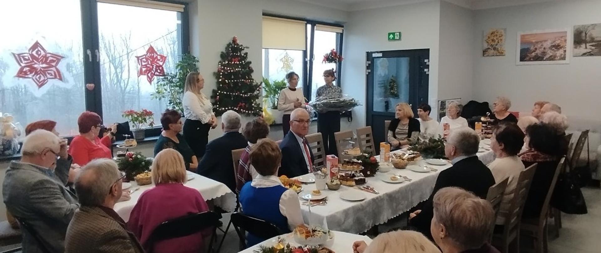 Przy świątecznie zastawionych stołach siedzą seniorzy i patrzą w kierunku stojących pod oknem i składających życzenia przedstawicieli gminy, na ścianie i oknach dekoracje świąteczne