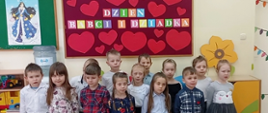 Zdjęcie przedstawia 13 dzieci w wieku 6 lat, ubranych w galowe stroje pozujących do zdjęcia na tle gazetki na której widnieje napis Dzień Babci i Dziadka. Nad napisem i pod napisem przyklejone są czerwone serduszka różnej wielkości. Miejsce zdjęcia to sala szkolna.