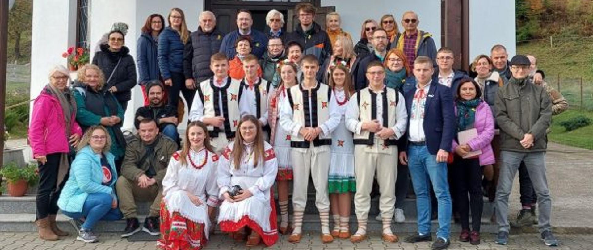 Grupa ludzi w ludowych strojach oraz kurtkach stoi na tle białego budynku w górach 