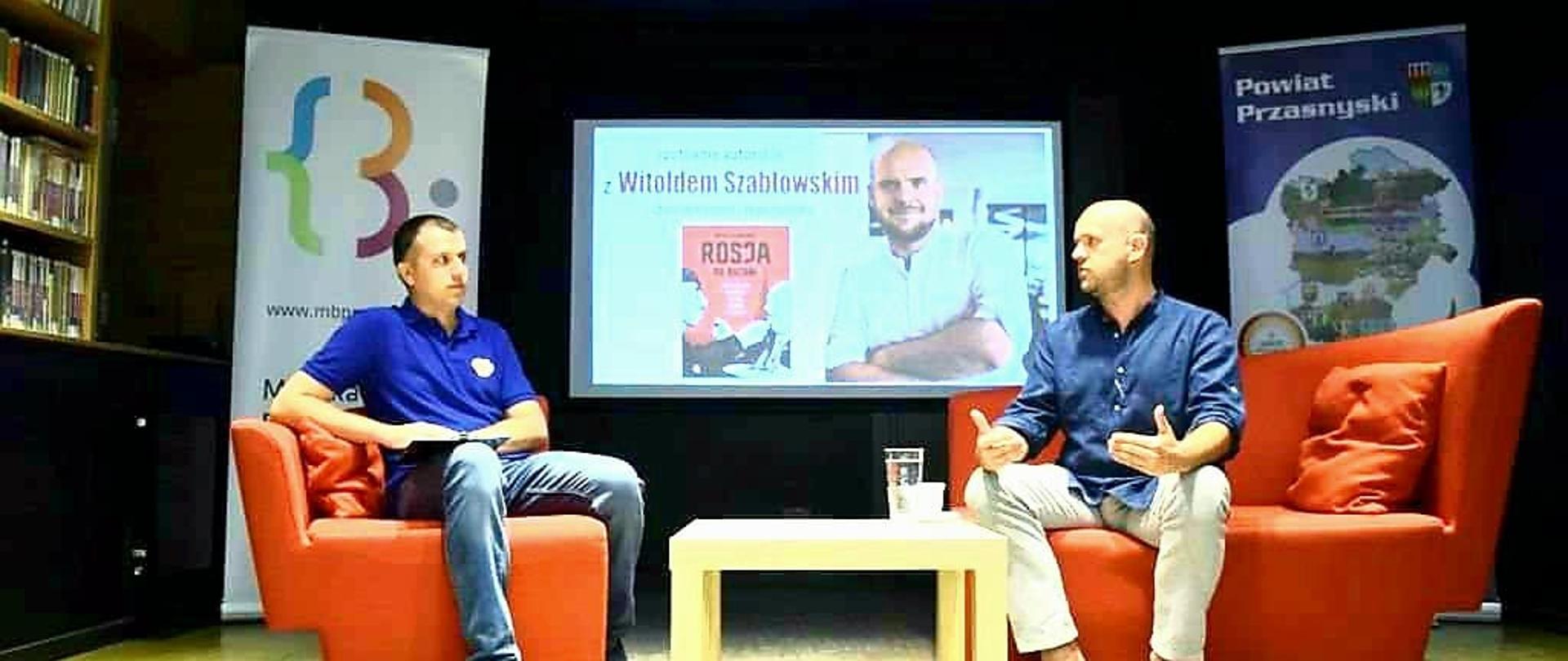 Zdjęcie wykonane wewnątrz Miejskiej Biblioteki Publicznej podczas spotkania z autorem Witoldem Szabłowskim. Na zdjęciu od prawej: Witold Szabłowski, pracownik biblioteki prowadzący spotkanie.