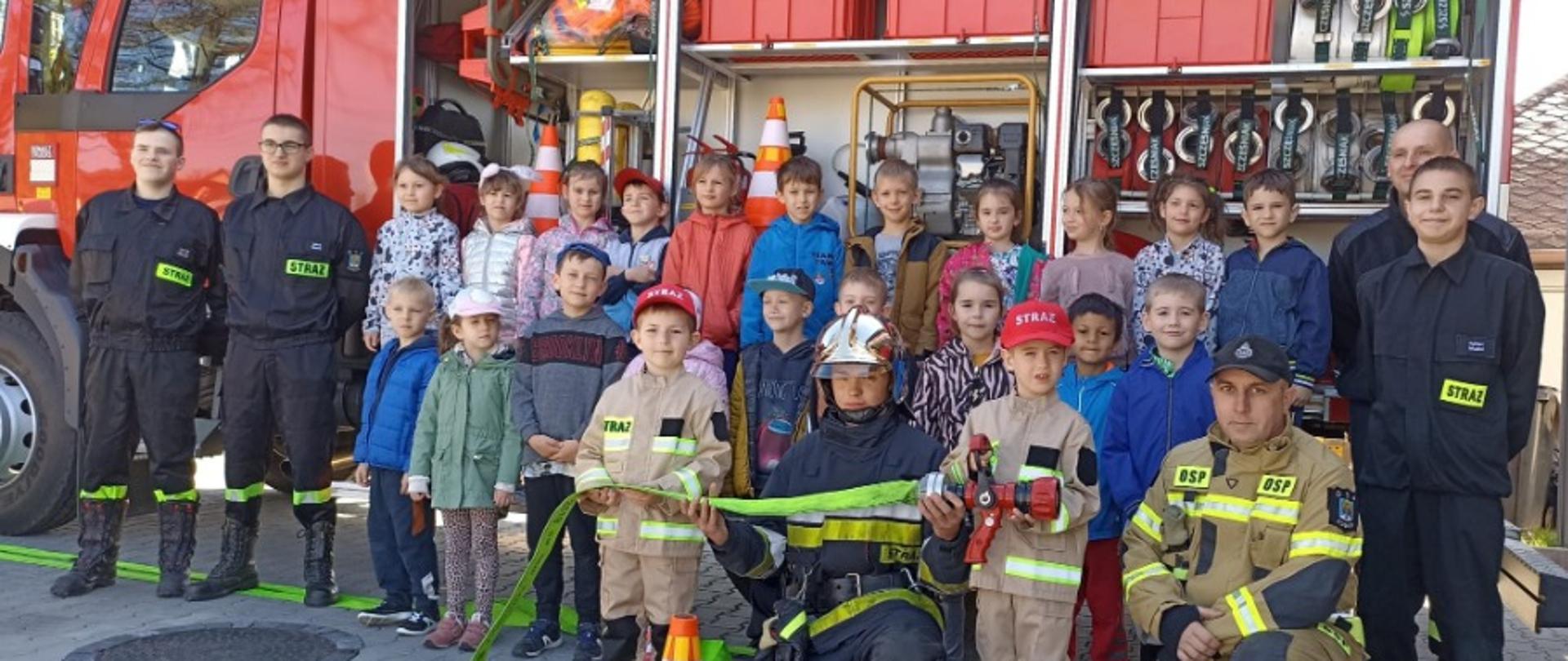 Grupa dzieci z przedszkola razem z kilkoma strażakami z OSP Kobiór na tle otwartego wozu strażackiego z wyposażeniem