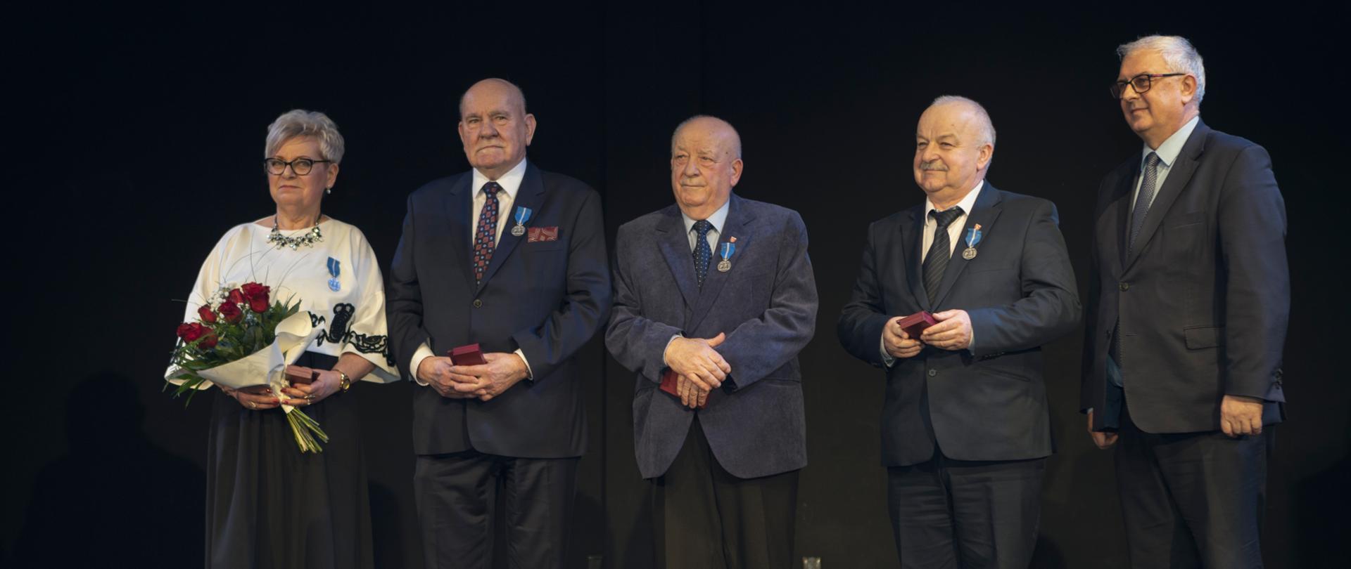 Pięć osób elegancko ubranych, czterech mężczyzn oraz jedna kobieta. Kobieta trzyma bukiet kwiatów.