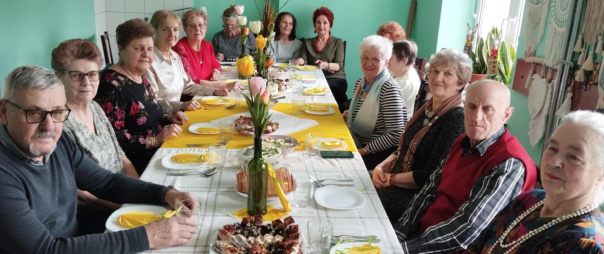 Członkowie klubu senior+ siedzący przy wielkanocnym stole. Stół przyozdobiony tulipanami, na stole stojące wielkanocne potrawy i ciasto.