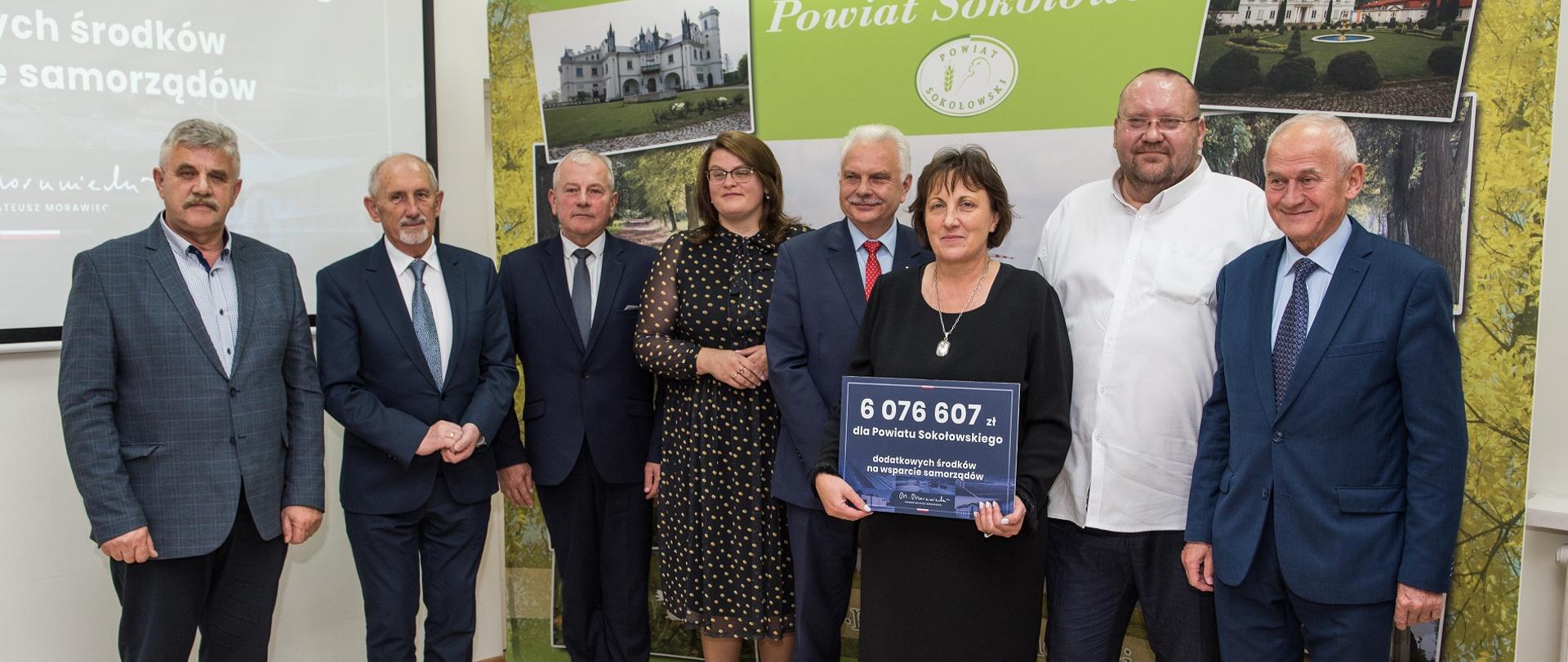 Zdjęcie grupowe reprezentacji Powiatu Sokołowskiego z wręczającymi czeki przedstawicielami rządu RP. W tle ścianka promocyjna Powiatu Sokołowskiego oraz slajd informacyjny o dofinansowaniu dla Powiatu.