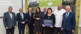 Zdjęcie grupowe reprezentacji Powiatu Sokołowskiego z wręczającymi czeki przedstawicielami rządu RP. W tle ścianka promocyjna Powiatu Sokołowskiego oraz slajd informacyjny o dofinansowaniu dla Powiatu.