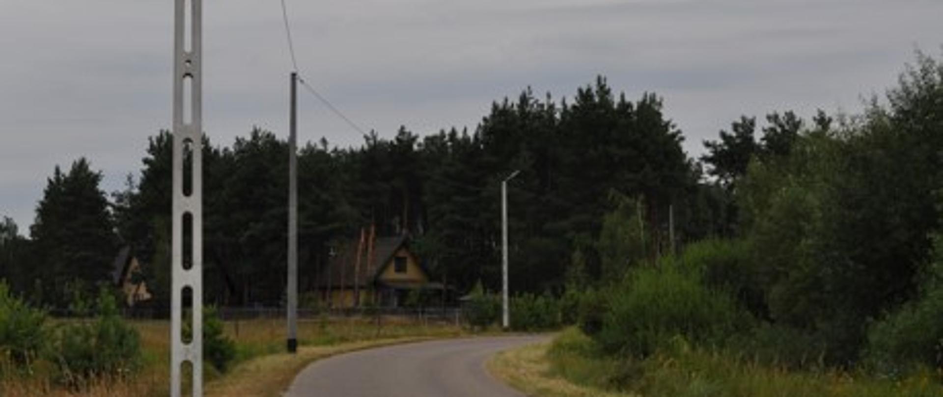 Na zdjęciu znajduje się nowo wybudowany odcinek oświetlenia ulicznego na ul. Nakieł w Brańszczyku. Po obu stronach drogi rosną trzewa i krzewy. W oddali znajdują się dwa domy.