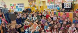 Dzieci siedzą na podłodze w sali przedszkolnej, słuchają pogadanki pani pedagog Małgorzaty Młotkowskiej na temat bezpiecznego wypoczynku podczas ferii, w tle tablica interaktywna, po lewej i prawej stronie widoczne szafki przedszkolne oraz zabawki