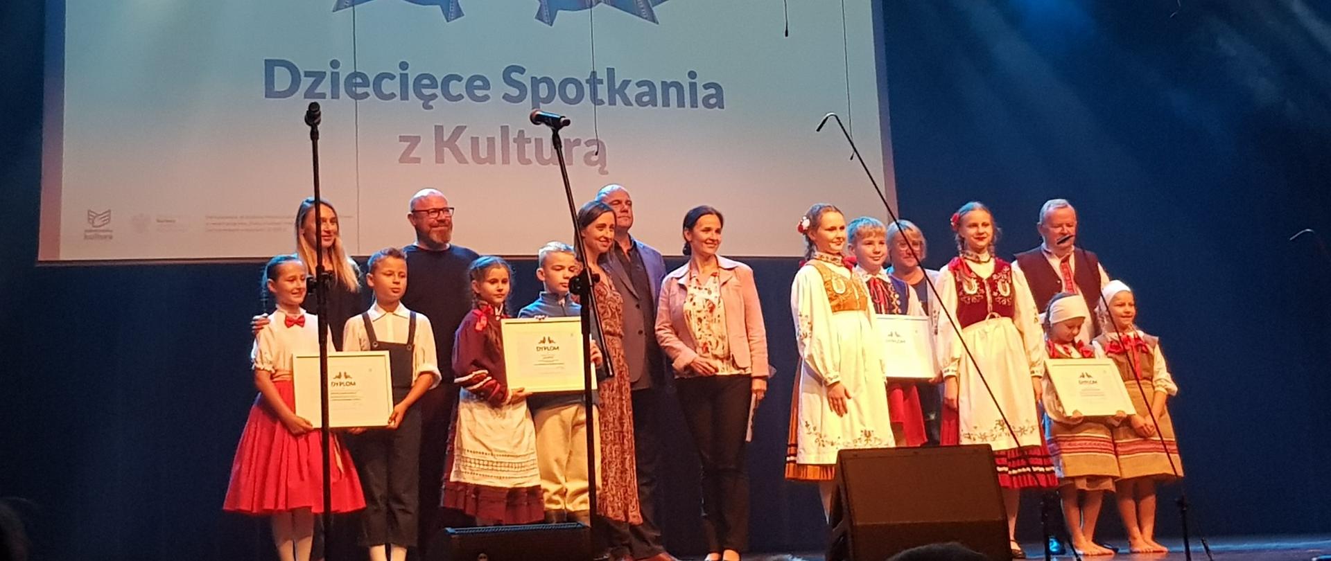 Dzieci z wychowawcami na scenie na tle napisu Dziecięce Spotkania z Kulturą