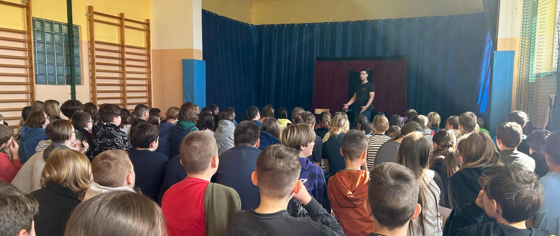 Mężczyzna w czarnej koszulce trzyma obie ręce przed sobą. Na przeciwko niego siedzi cała sala dzieci