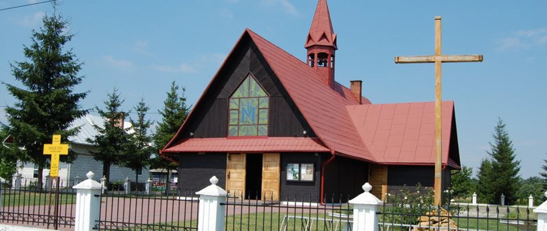 Drewniany, brązowy kościół z czerwonym dachem i wieżyczką. Z przodu budynku duże witrażowe okno. Przed kościołem drewniany krzyż. 