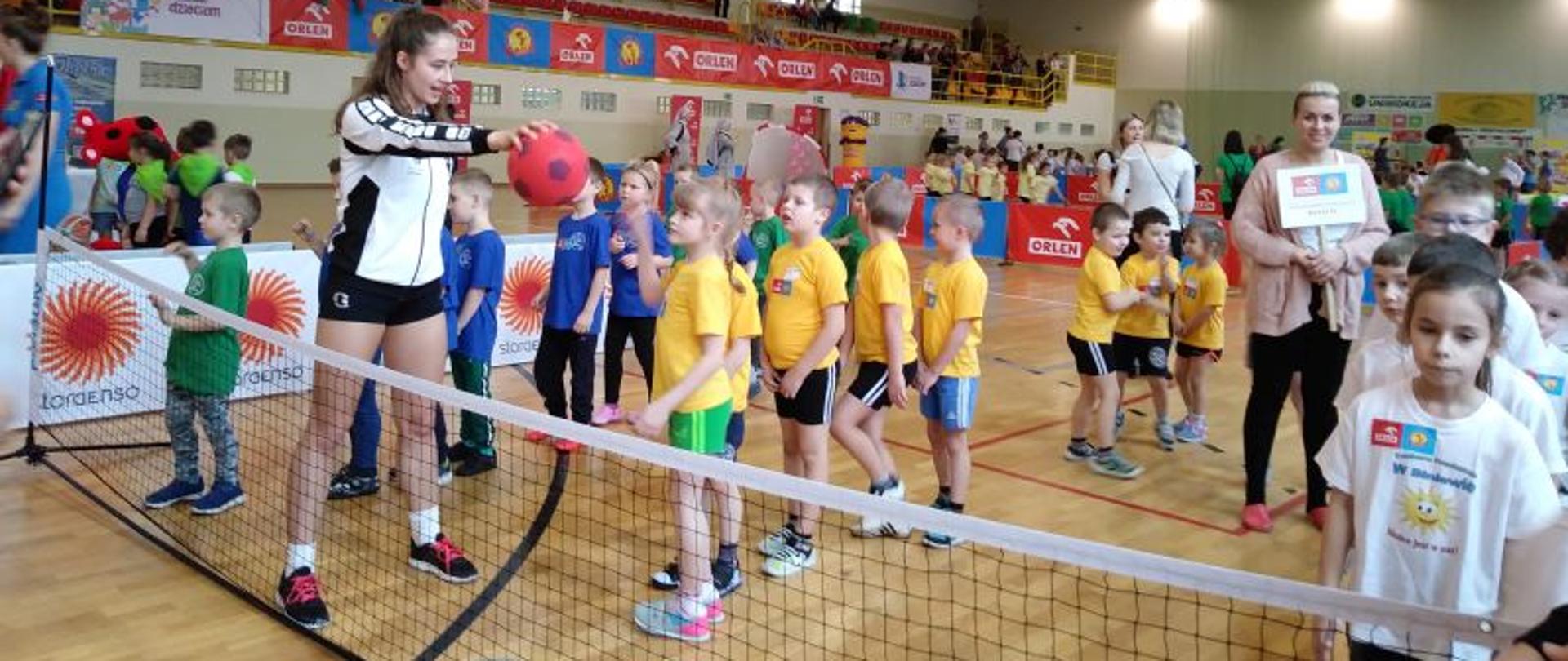 Trzy grupy dzieci w różnokolorowych koszulkach stojące w rzędach przed siatką, młoda dziewczyna podaje piłkę, inna trzyma w rękach plakat. W tle banery, widzowie i inne grupy dzieci. 