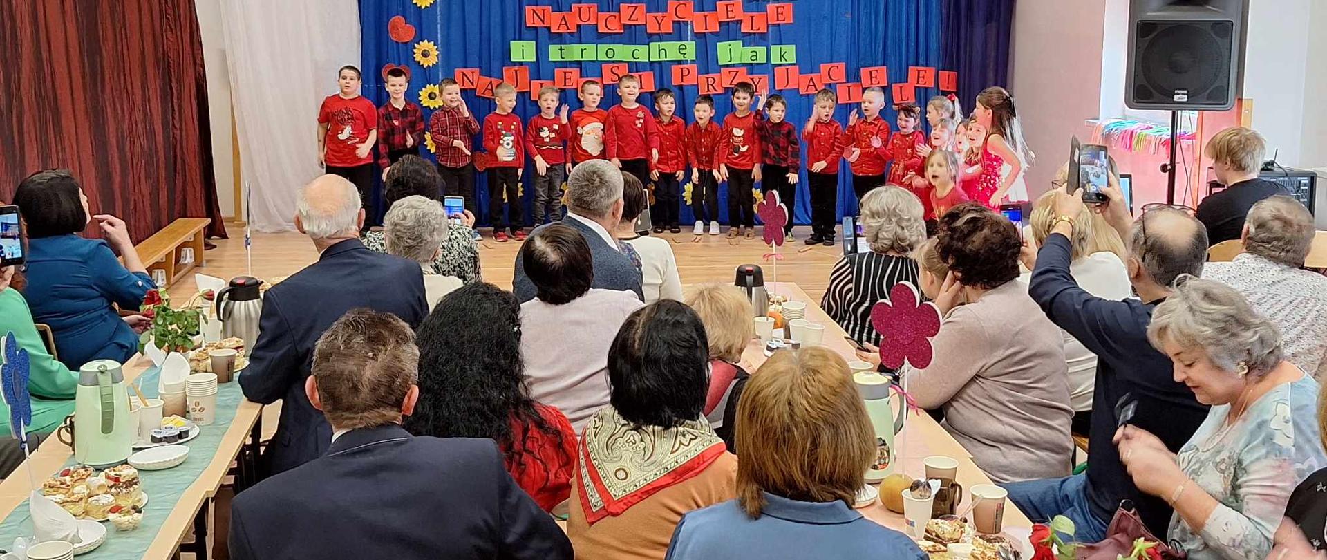 Dzieci ubrane w czerwone stroje śpiewają piosenkę. Wokół siedzą zebrani goście.