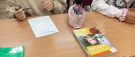 Na szkolnej ławce leży karta do głosowania, dwa podręczniki i piórnik z przyborami piśmiennymi, widać ręce siedzącej w ławce uczennicy