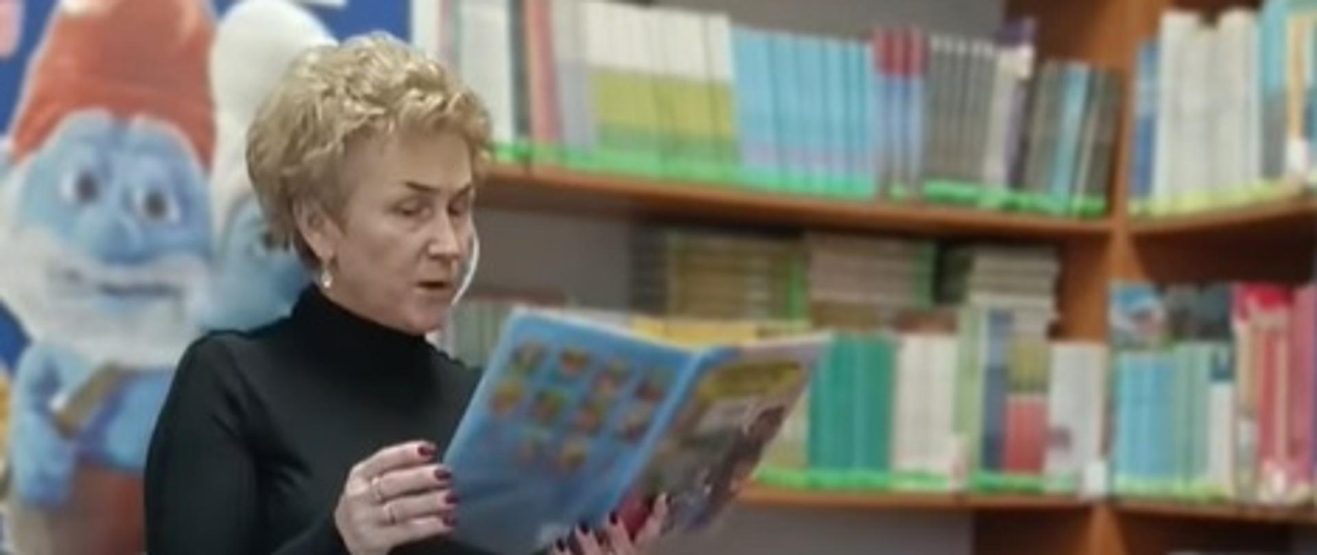 Bibliotekarka czyta przedszkolakom jedną z nowo zakupionych książek, w tle dekoracja z postaciami z bajek i regały z księgozbiorem
