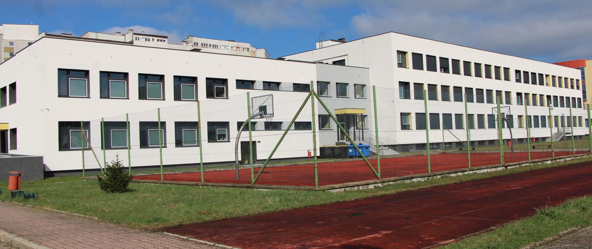 Wielopiętrowy budynek, z białą elewacją , na pierwszym planie obiekty sportowe z boiskiem do koszykówki i bieżnią do biegania.