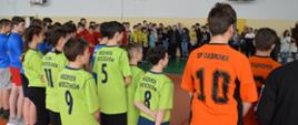 Zdjęcie przedstawia dużą grupę młodych zawodników, zarówno chłopców, jak i dziewcząt, którzy ubrani w stroje swoich reprezentacji szkolnych stoją ustawieni w okręgu dookoła hali sportowej