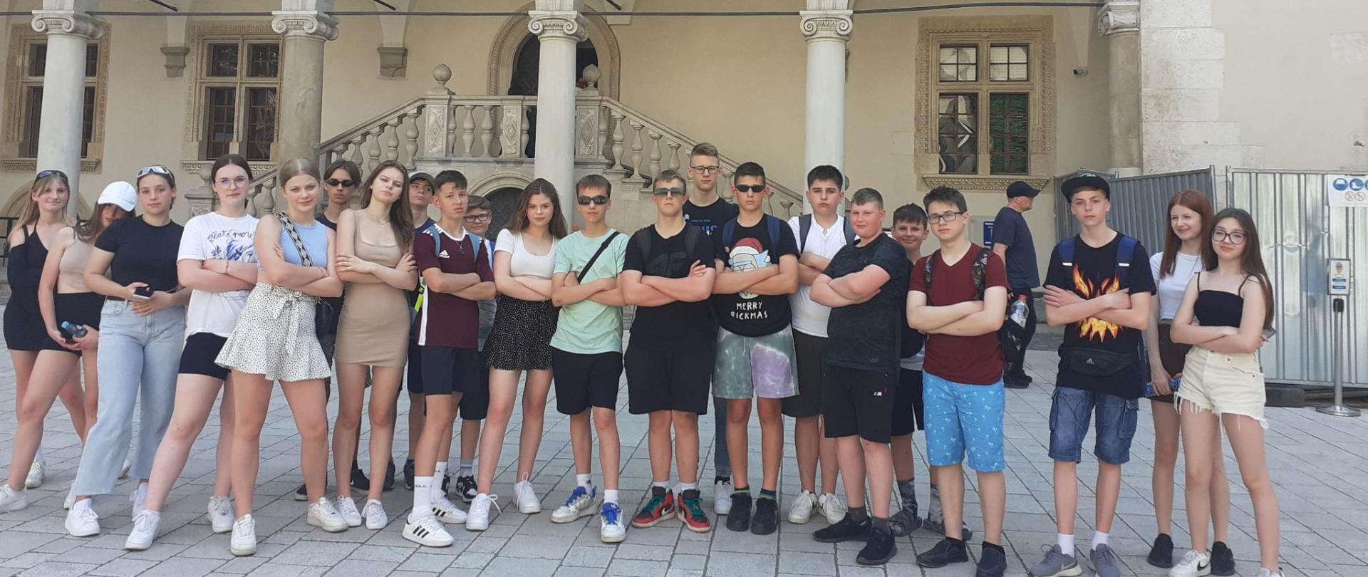 Uczniowie klasy VIII ustawieni na tle architektury krakowskiego rynku z założonymi rękami spoglądają w kierunku obiektywu aparatu.
