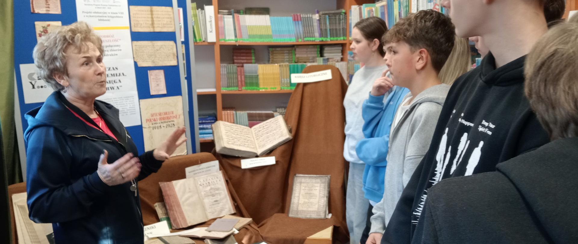 Bibliotekarka pokazuje zgromadzonym uczniom stare grube księgi, w tle regały biblioteczne