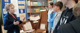 Bibliotekarka pokazuje zgromadzonym uczniom stare grube księgi, w tle regały biblioteczne