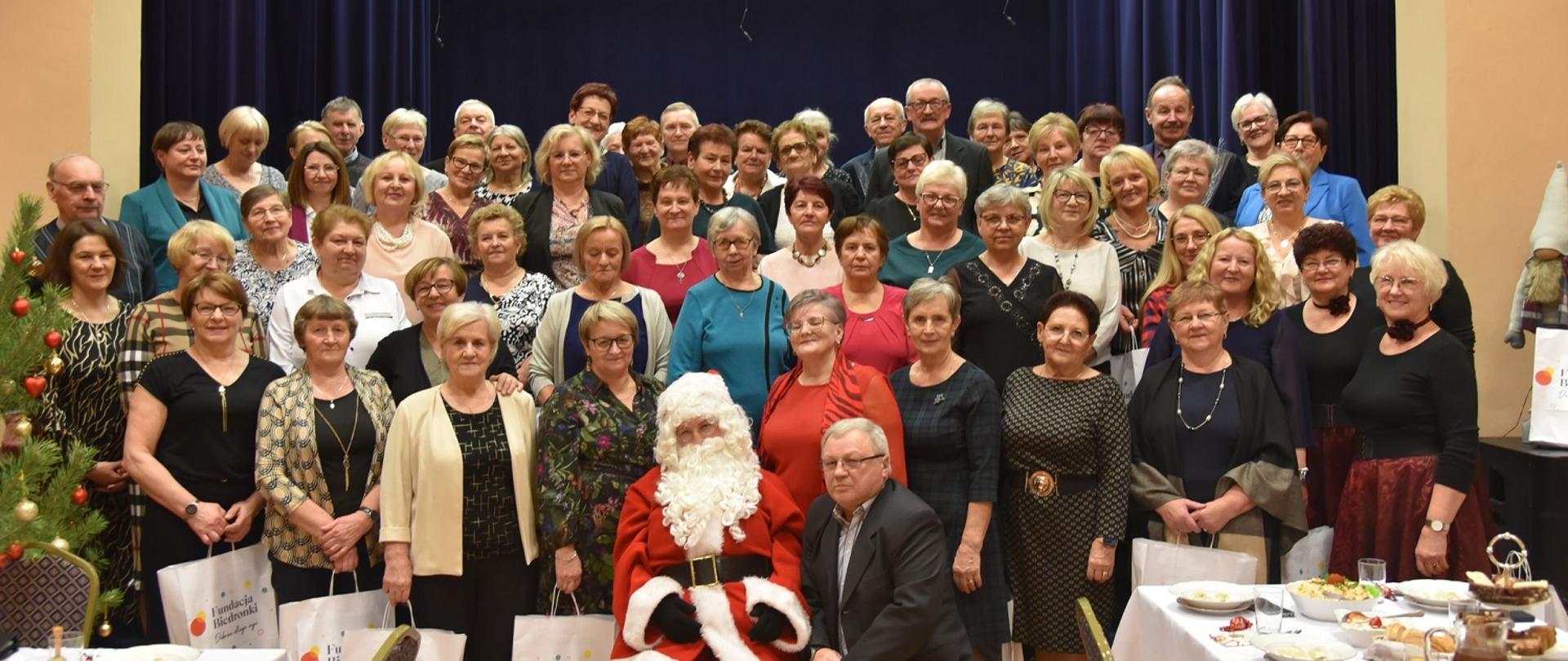 kobiety i mężczyźni w odświętnych strojach pozują do zdjęcia, na środku siedzi św. Mikołaj 