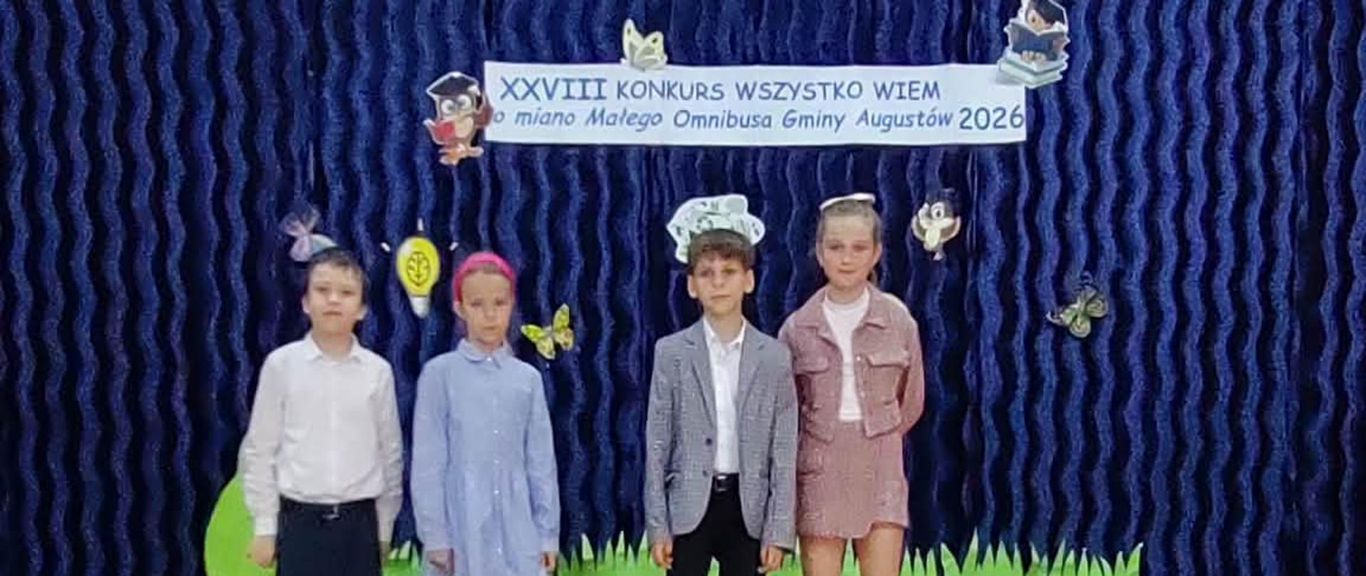 Na scenie stoi czterech reprezentantów naszej szkoły w konkursie wiedzy „Wszystko wiem o miano Małego Omnibusa Gminy Augustów” - Michał Wróblewski z Oddziału Przedszkolnego A, Aleksandra Tężycka z klasy pierwszej, Antonina Moroz z klasy drugiej i Dominik Burak z klasy trzeciej. Wszyscy ubrani są odświętnie. 