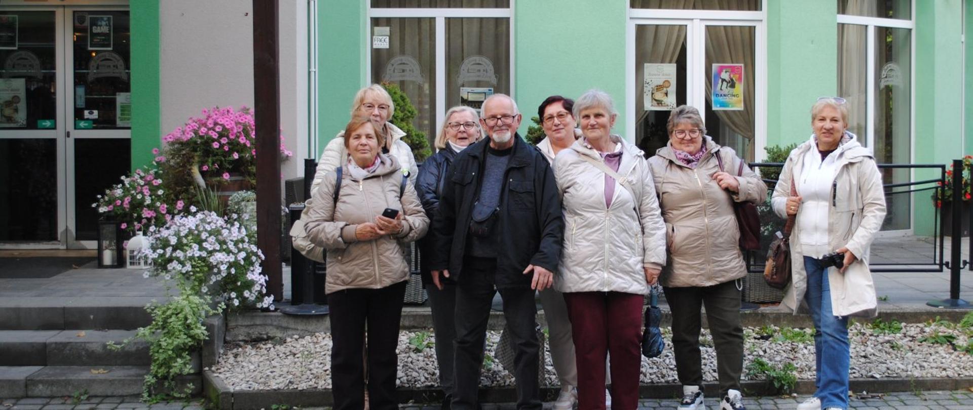 siedem kobiet i jedne mężczyzna stojący w środku pozują do zdjęcia na tle zielonego budynku 