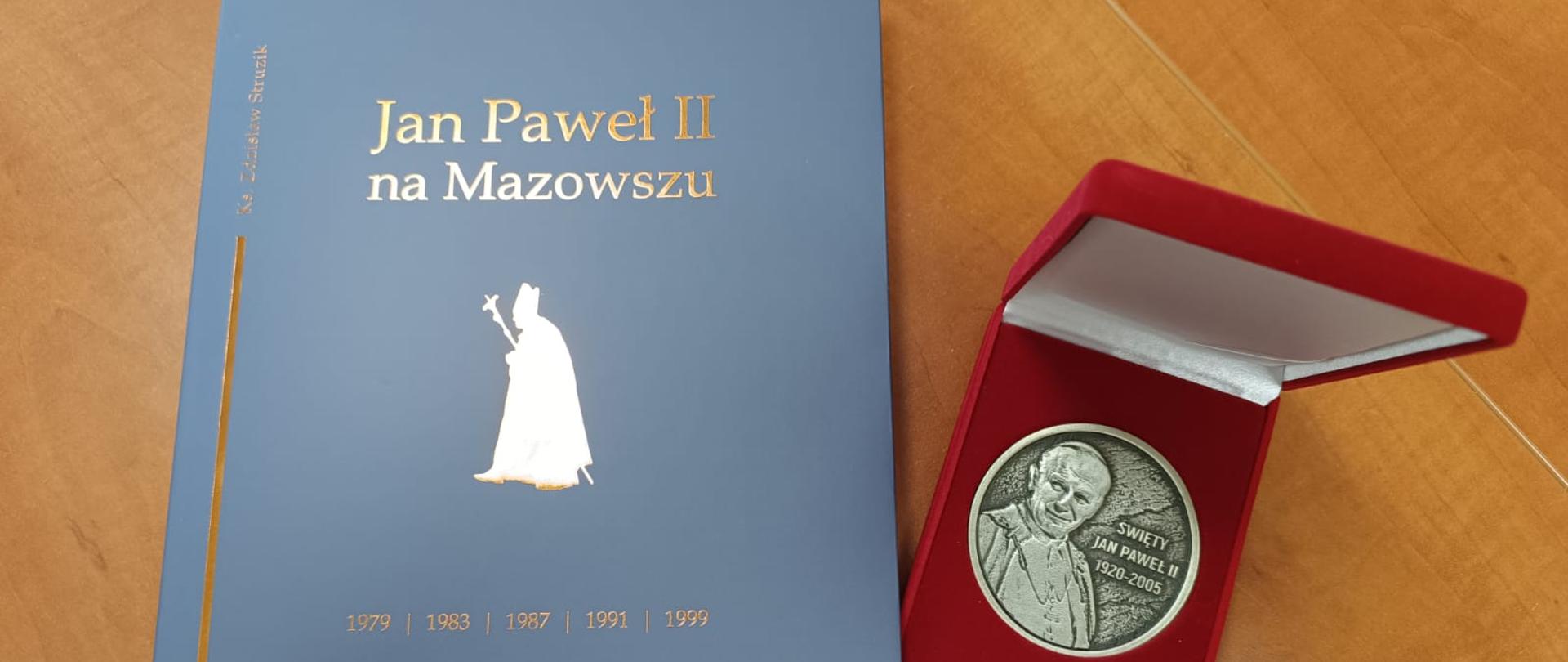 Zdjęcie przedstawia medal okolicznościowy z okazji 20-lecia Instytutu Papieża Jana Pawła II oraz publikację jubileuszową "Jan Paweł II na Mazowszu".