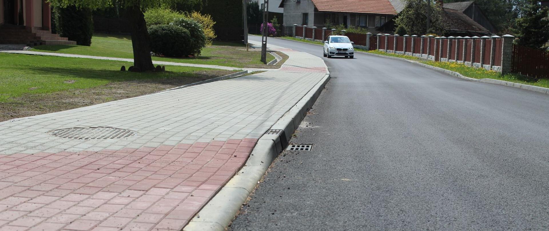 Droga gminna w Stanisławiu Dolnym po przeprowadzonej modernizacji. W centralnym miejscu drga asfaltowa, po lewej chodnik z kostki brukowej, w oddali samochód i zabudowania.