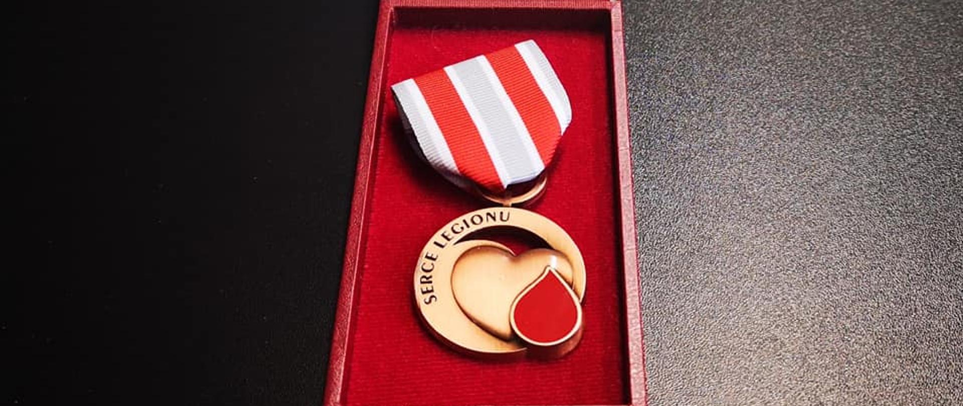 Zdjęcie przedstawia brązowe odznaczenie Honorowych Dawców Krwi Legion - serce z czerwoną kroplą krwi oraz napis "Serce Legionu". Odznaka leży na czerwonym materiale w drewnianym obitym na czerwono futerale.