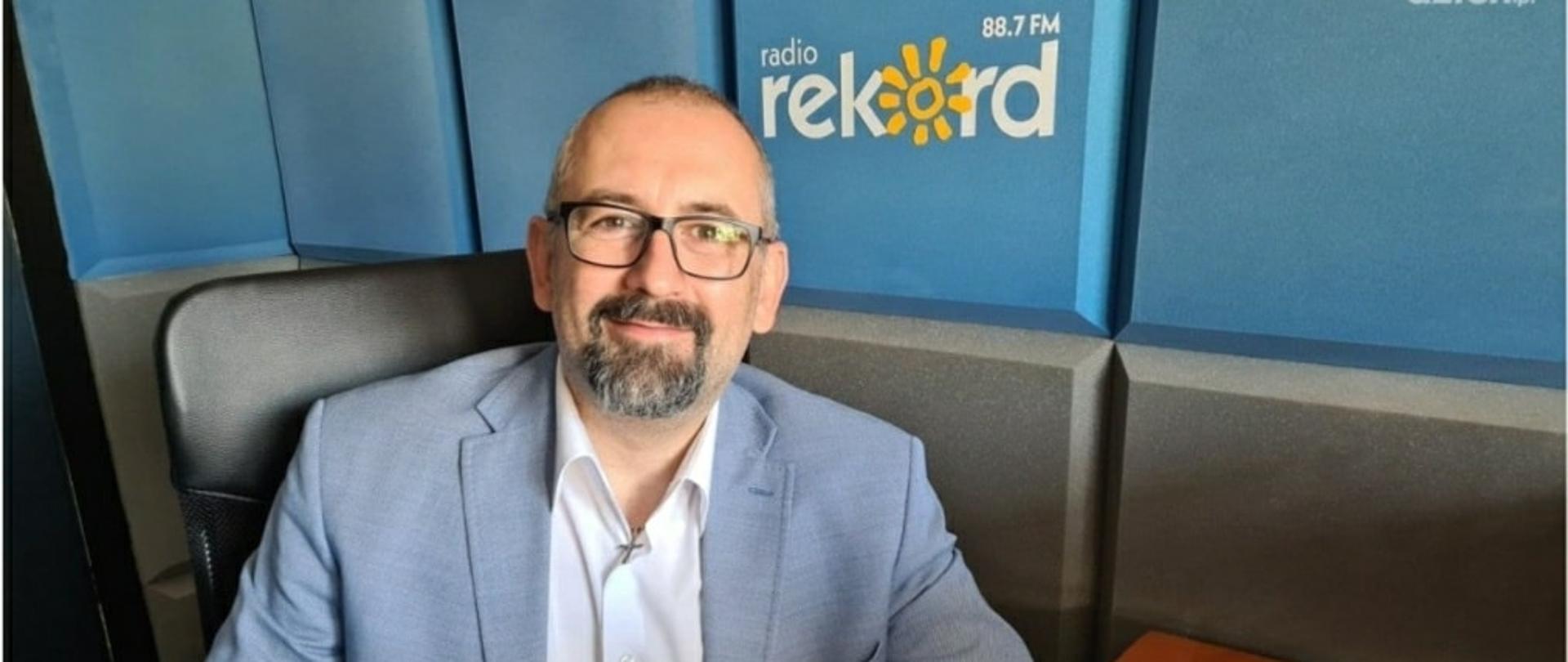 Starosta Przasnyski Krzysztof Bieńkowski siedzi uśmiechnięty w błękitno szarym studiu Radia Rekord 88,7 FM w Ciechanowie.