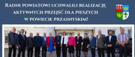 Grafika upamiętniająca wydarzenie uchwalenia głosami radnych Powiatu Przasnyskiego realizacji aktywnych przejść dla pieszych w powiecie przasnyskim. U góry biały napis o treści "Radni powiatowi uchwalili realizację aktywnych przejść dla pieszych w powiecie przasnyskim!", po prawej obok napisy herb Powiatu Przasnyskiego. Pod spodem na szerokości grafiki zdjęcie przedstawiające radnych powiatu powiatowych stojących w szeregu przed wejściem do budynku Starostwa Powiatowego w Przasnyszu. Pod spodem dwa zdjęcia, po prawej przejście dla pieszych, po lewej wyświetlacz prędkości zasilany panelem słonecznym.