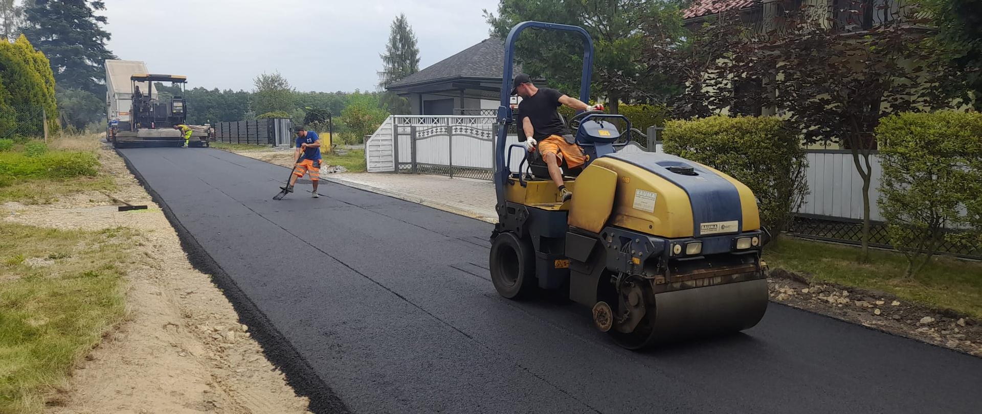 Walec drogowy ubija świeżo położony asfalt w tle domy i robotnicy poprawiający nawierzchnię